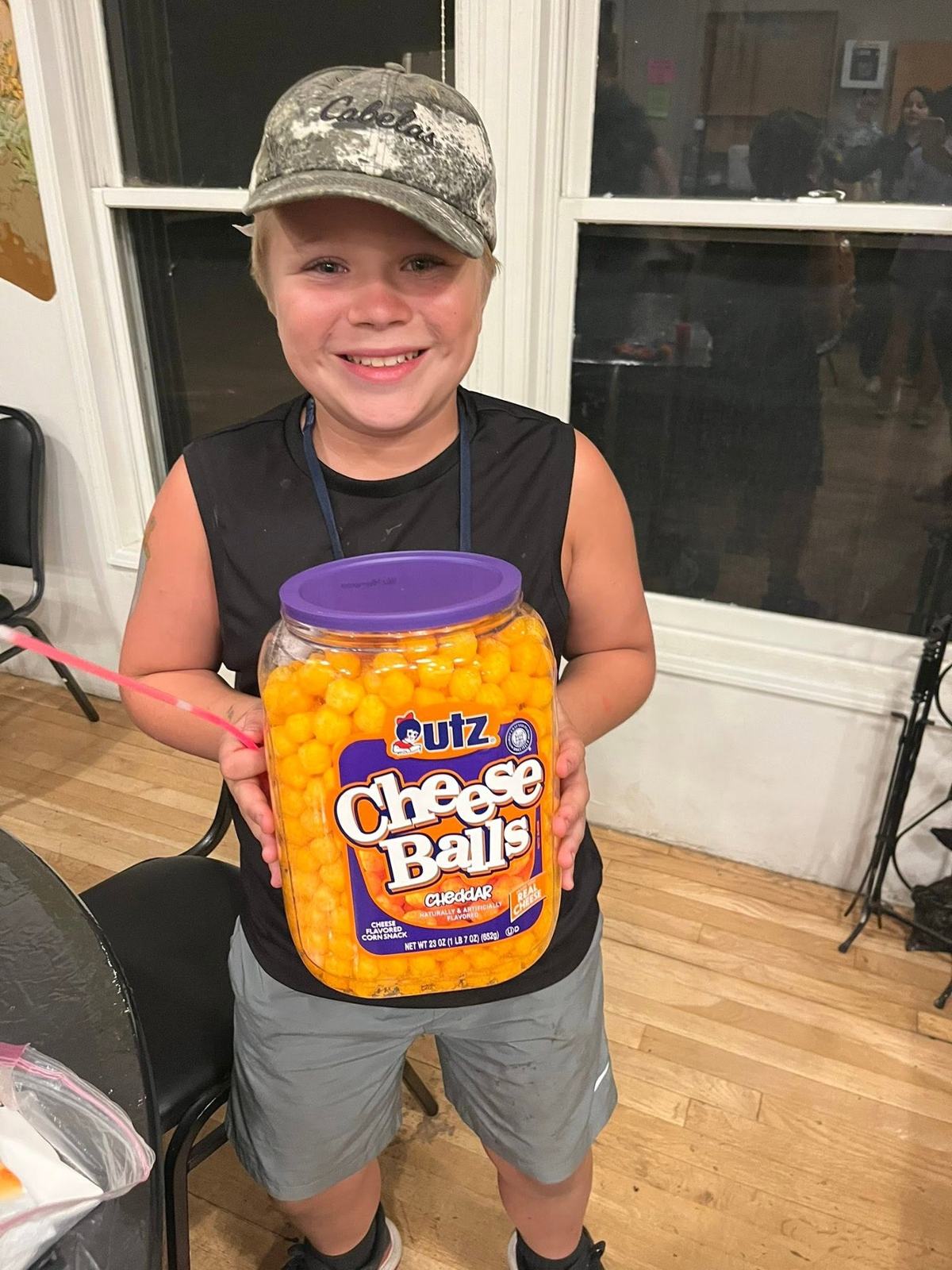 A grinning boy at camp proudly holding a giant tub of Utz cheese balls