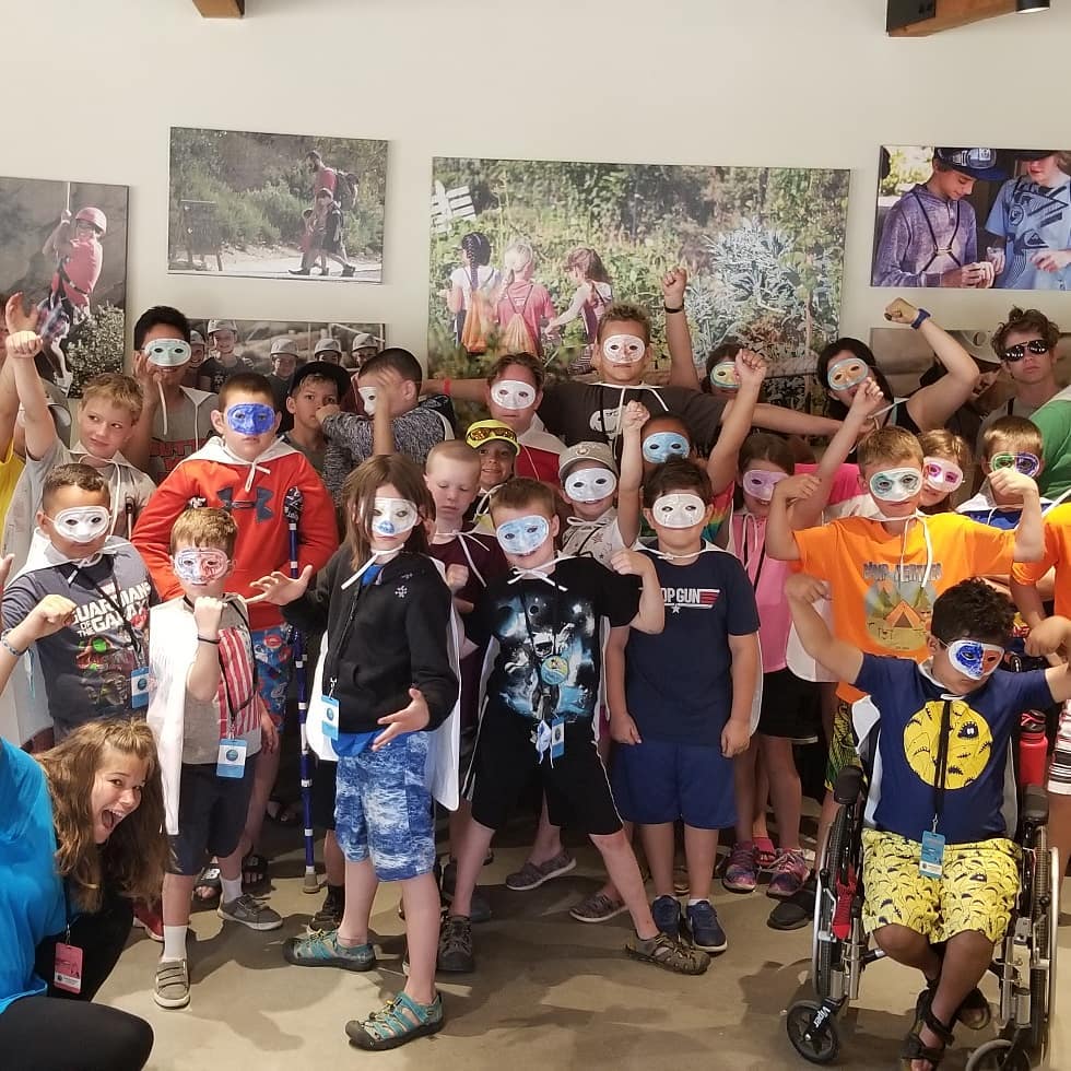 A whole camp of kids in white superhero masks flexing their muscles together, crutches and a wheelchair among them