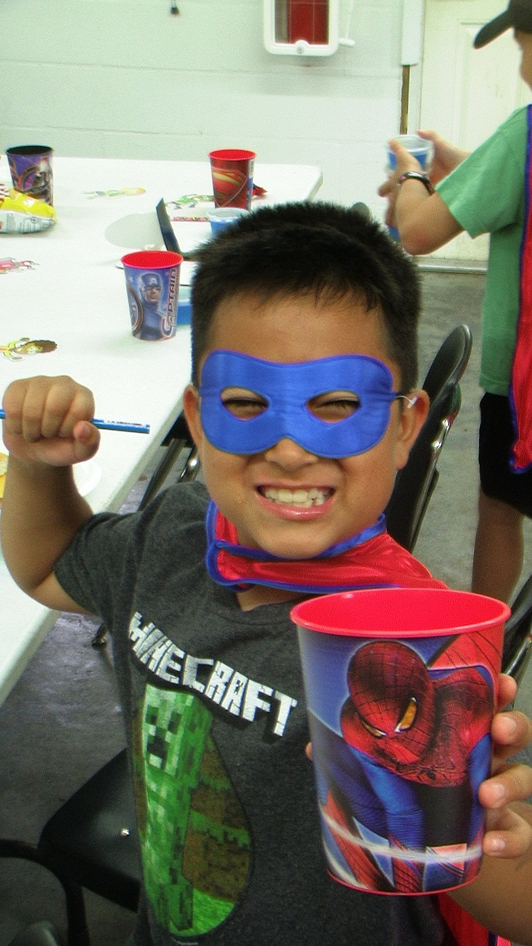 A boy in a blue superhero mask and cape grinning and raising his fist at a craft table