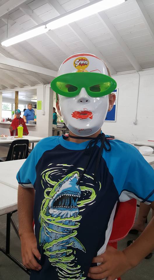 A camper close up in a white superhero mask with a drawn-on grin, green visor and red cape, hand on hip