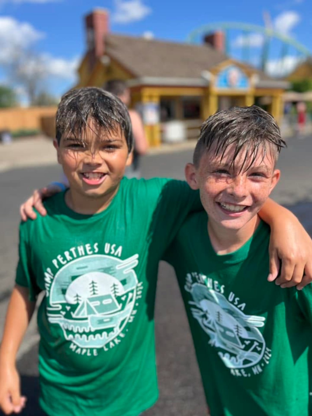 Two boys in green Camp Perthes USA shirts with arms around each other, grinning at an amusement park